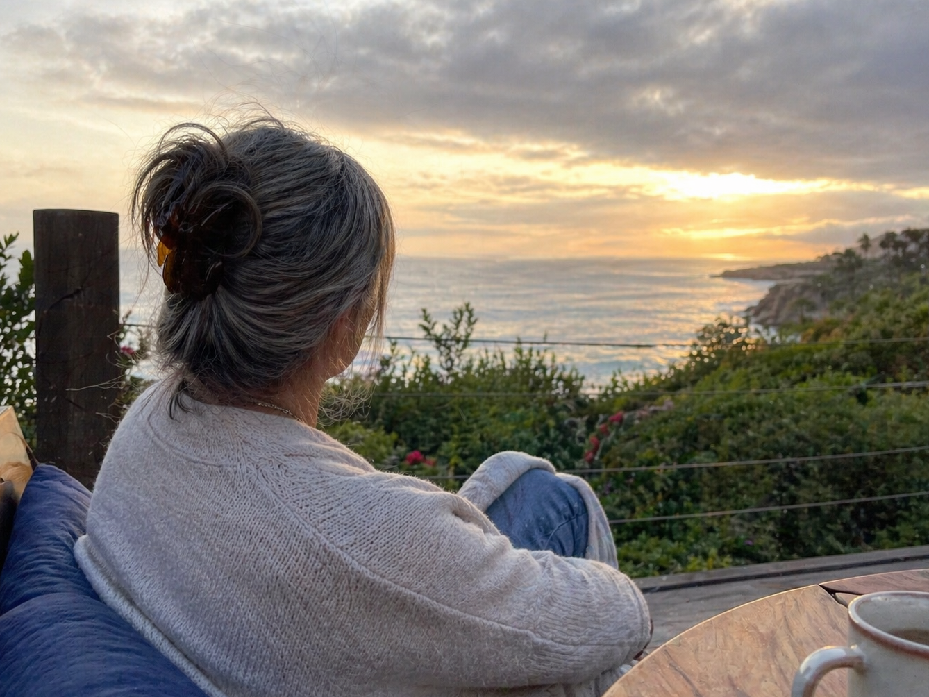 A woman with gray hair sitting on an outdoor deck, gazing at the sunset with a cup of coffee beside her.