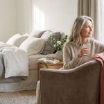 A mature woman drinks water beside a sunny window in a calm bedroom.