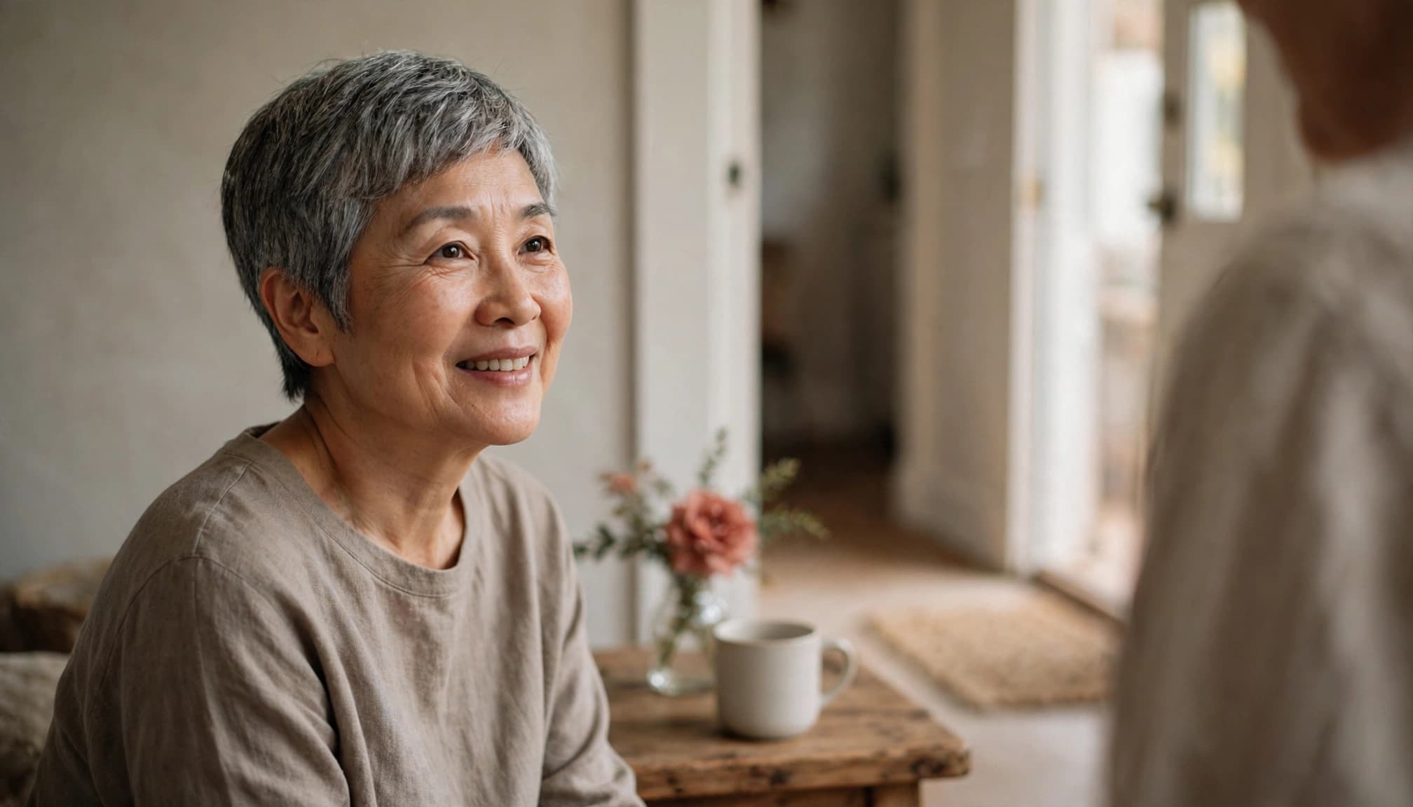 Older Asian woman speaking gently with a friend at a home doorway, beside a mug and coral flower.