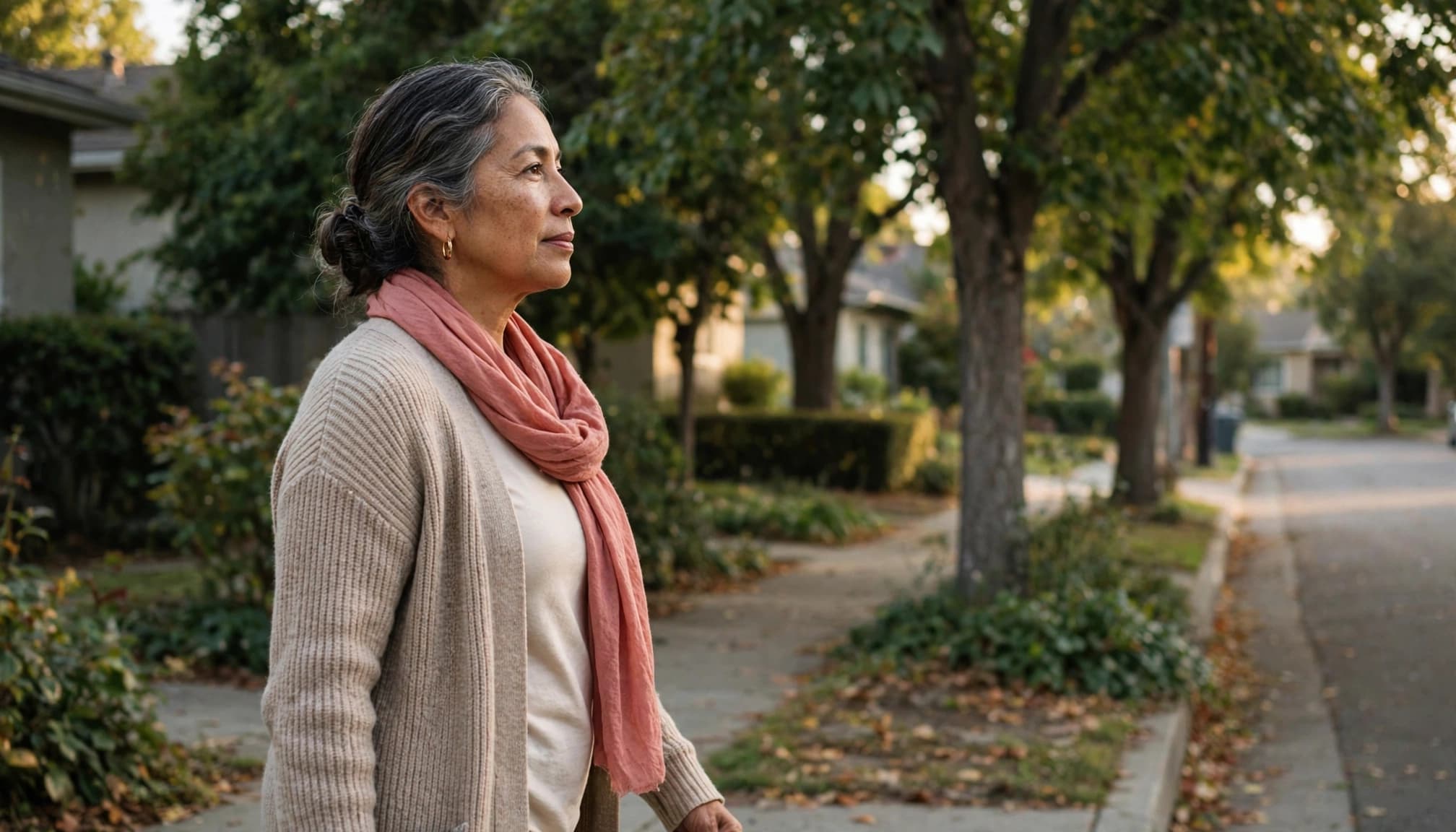 Older woman walking alone on a leafy sidewalk in warm afternoon light.